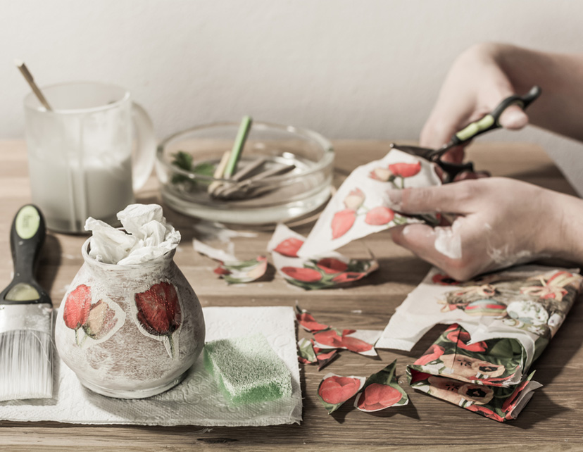 Decoupage accessories on a table - brush, scissors, paper, tissues, sponge, pencils, paint. Hands cutting flowers in the background.